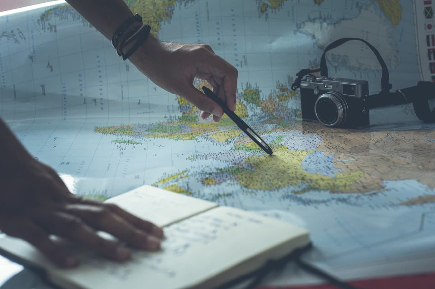 A hand tracing a route on a world map with a pen, beside a notebook holding a handwritten list.